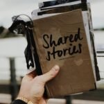 Hand holds a "Shared Stories" journal against a blurred outdoor backdrop, emphasizing creativity and personal storytelling.